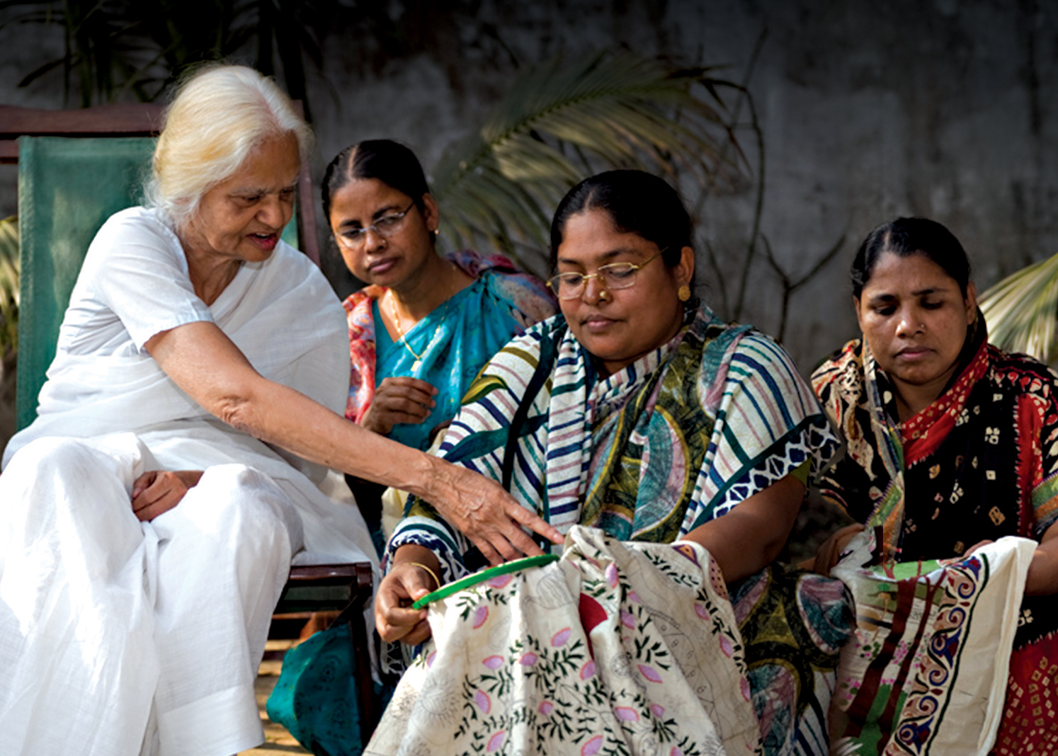 Image of Surayia Rahman with three fellow artisan embroiderers. One woman has an embroidery hoop and Surayia is seen guiding the embroiderer while the other two women watch.